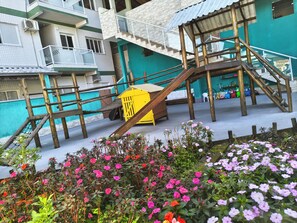 A multi-level wooden play structure with a yellow slide, surrounded by a garden with pink flowers. - Canto Verde Aptos II (Caraguatatuba)