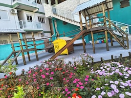 Uma estrutura de madeira com vários níveis e um escorregador amarelo, cercada por um jardim com flores cor-de-rosa.