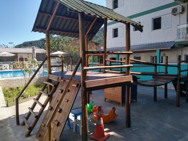 Eine Holzspielstruktur mit Rutschen und Kletterelementen, ein Schwimmbecken und ein Gebäude mit Kachelfassade.
