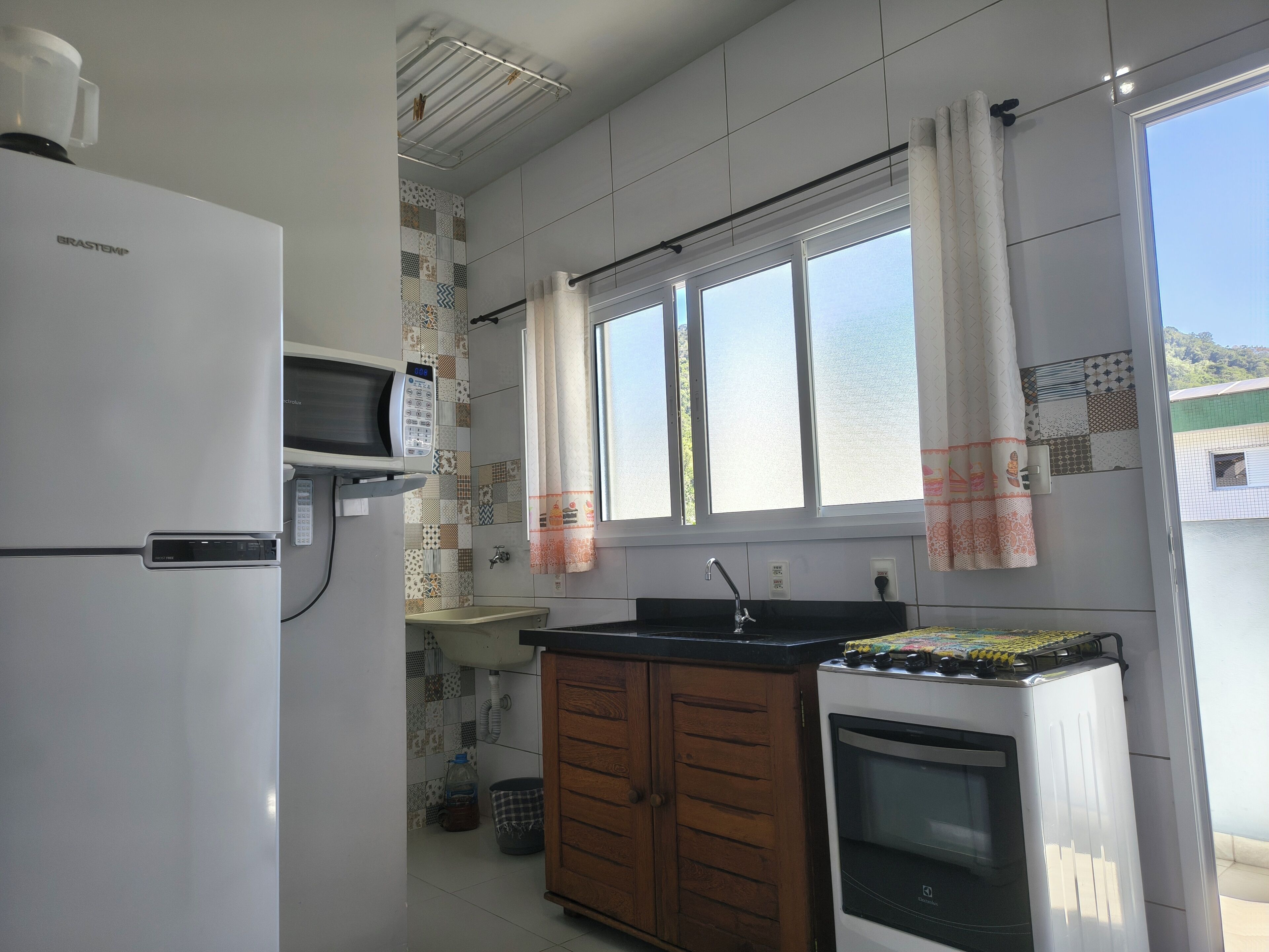 A kitchen with a refrigerator, microwave, sink, wooden cabinets, and a window with curtains.