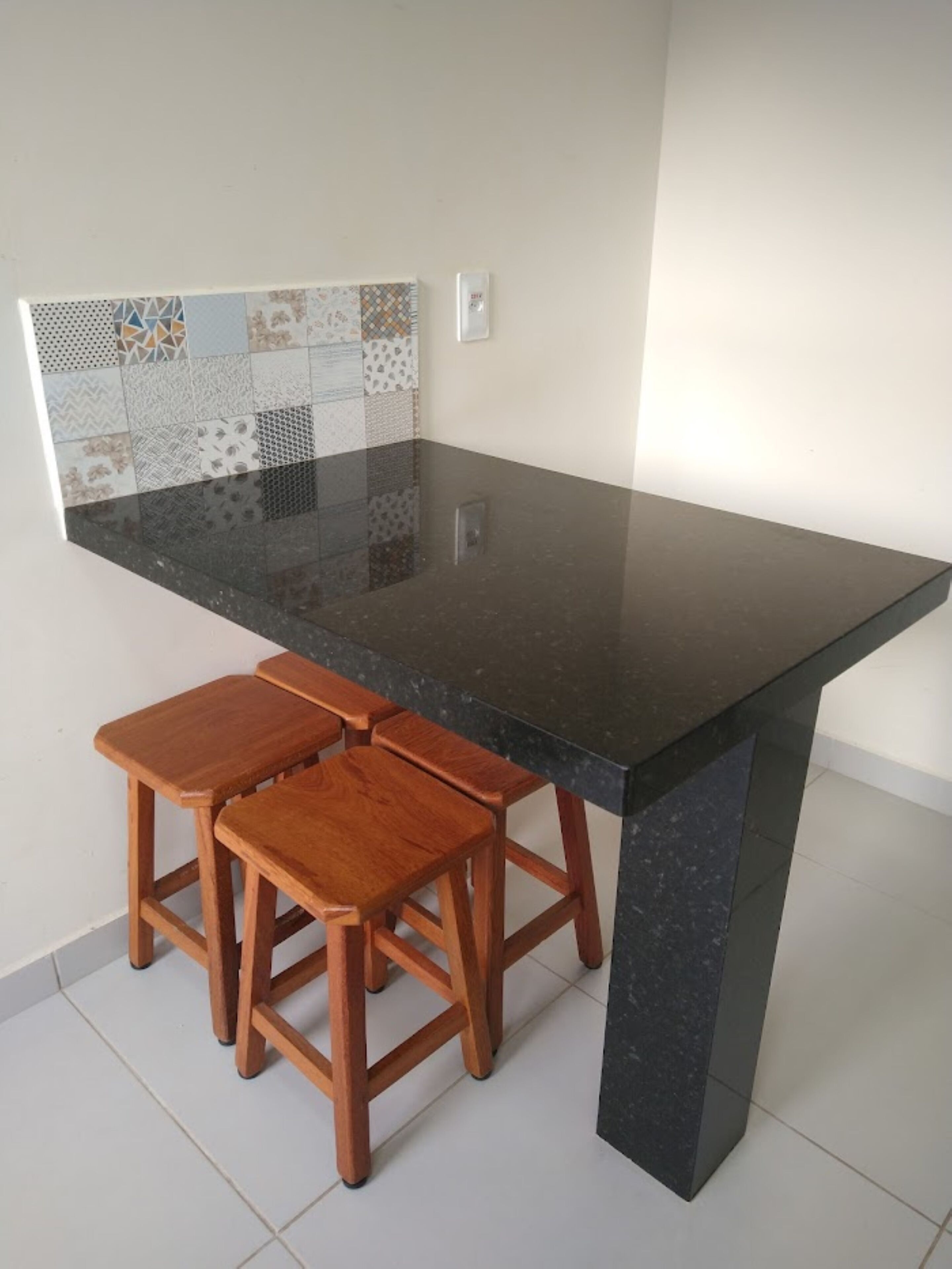 Une cuisine avec un plan de travail noir, des tabourets en bois et un mur recouvert de carreaux.