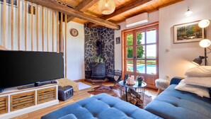 A cozy living room with a large flat-screen TV, a stone fireplace, and a view of a swimming pool. - Lovely home in Starcevljani (Starcevljani)
