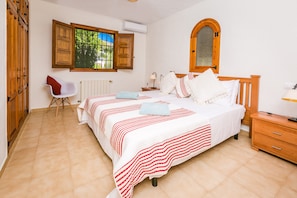 A bedroom with a large bed, a wooden wardrobe, a chair, and a window with open shutters.