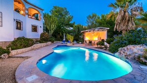 A swimming pool with a stone deck, surrounded by lush greenery and a patio area with outdoor seating.