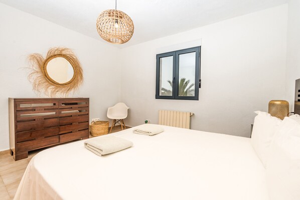 Een slaapkamer met een bed, een houten dressoir, een rieten lamp, een spiegel en een raam.
