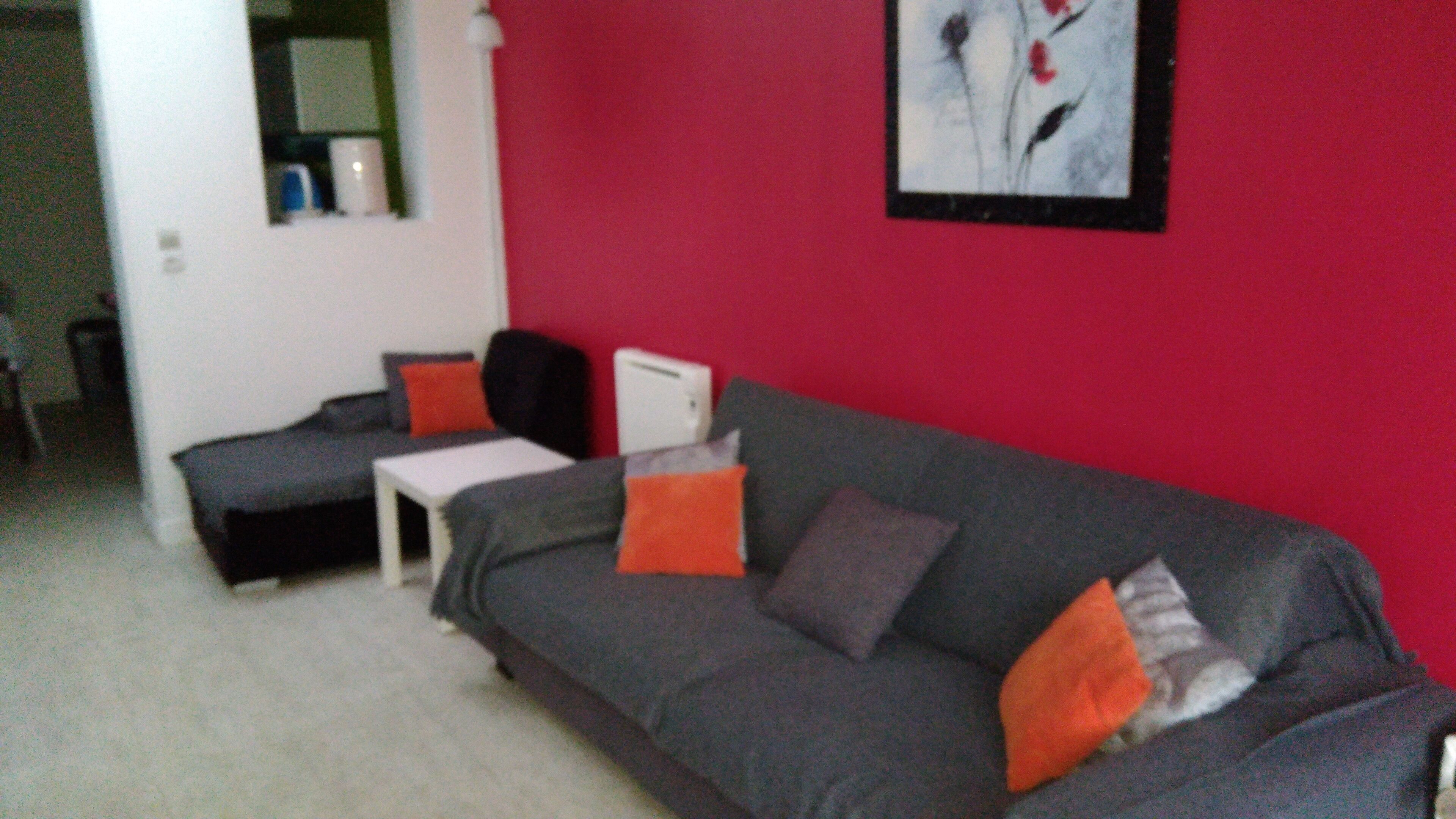 Un salón con un sofá gris, dos cojines anaranjados, una mesita de noche blanca y una pared roja.