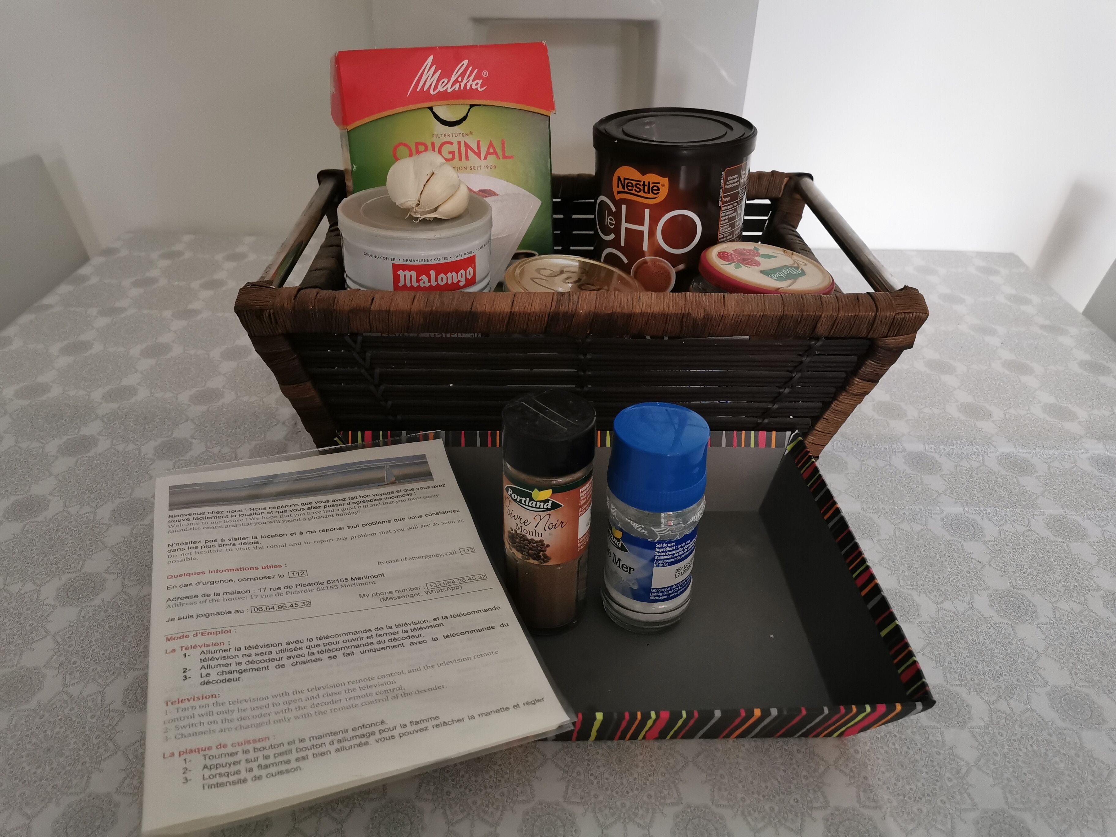 Un panier en osier contenant une boîte de café Melitta Original, un pot de pâte à tartiner au chocolat Nestlé, une bouteille de cognac Moutai, un paquet de café Portland et un menu.