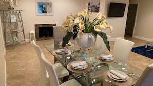 Een eetruimte met een glazen tafel voor zes personen, witte stoelen en een vaas met witte bloemen.