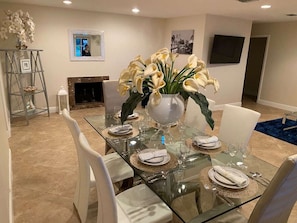 Uma sala de jantar com mesa de vidro posta para seis pessoas, cadeiras brancas e um vaso de flores brancas.