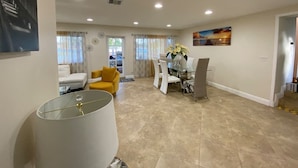 Een ruime woonkamer met een gele bank, een glazen salontafel en een eethoek met witte stoelen en een glazen tafel.