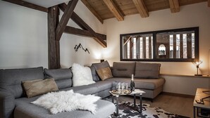 A cozy living room with a grey sofa, a white furry throw, and a glass table with a bottle and glasses. There's a window with a view of a building and a cowhide rug on the floor. - Nouveau Duplex Centre Village vue sur L'eglise et Terrasse sud (La Clusaz)