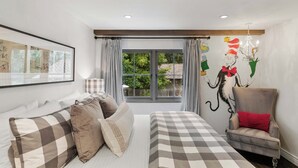 Un dormitorio con cama, sofá, sillón, ventana con cortinas y una pared con mural.