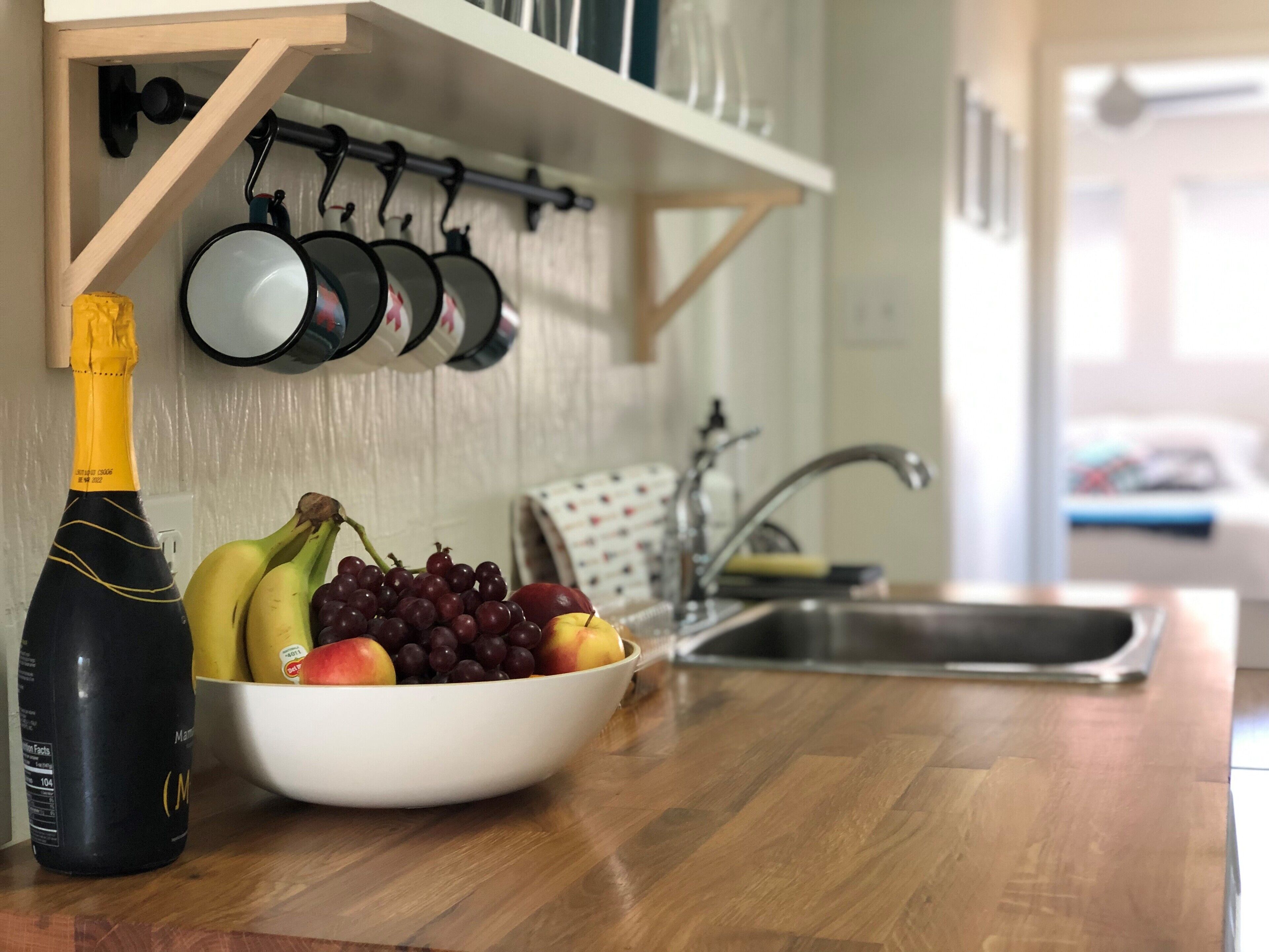 Una encimera de cocina con un cuenco de fruta, una botella de champán y ollas colgando.