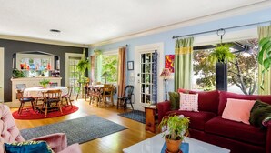 Ein geräumiges Wohnzimmer mit einem roten Sofa, einem Essbereich mit Tisch und Stühlen und großen Fenstern mit Blick auf viel Grün.