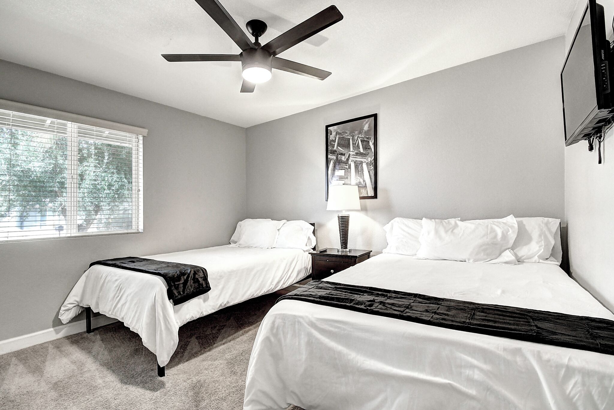 A hotel room with two beds, a ceiling fan, a window with blinds, and a framed picture on the wall.