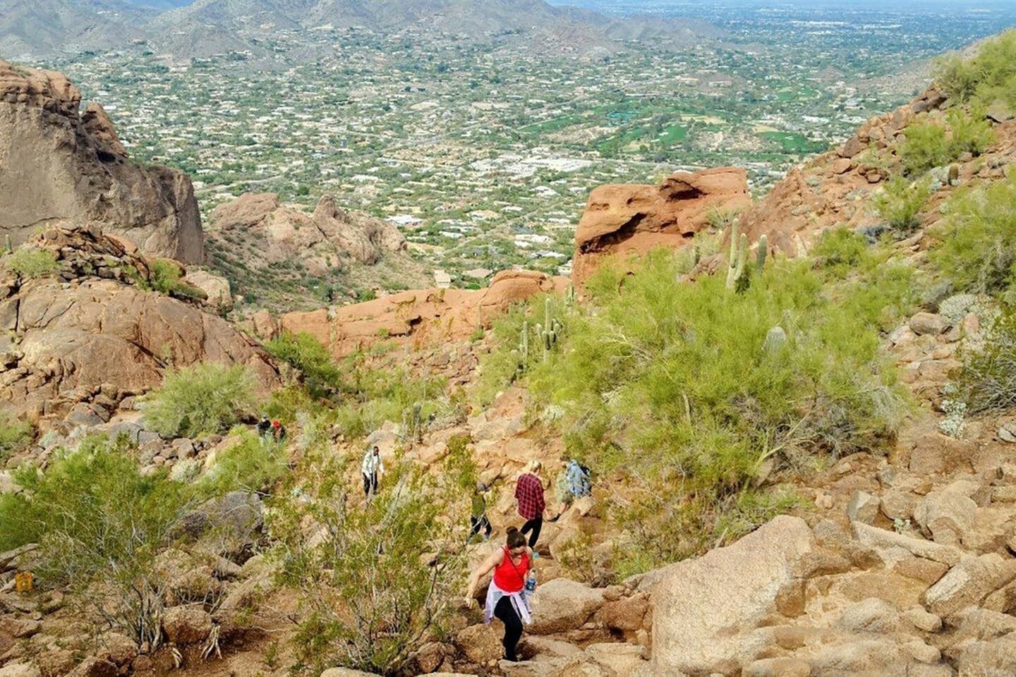 A rocky hiking trail with hikers, surrounded by sparse vegetation and a distant cityscape.