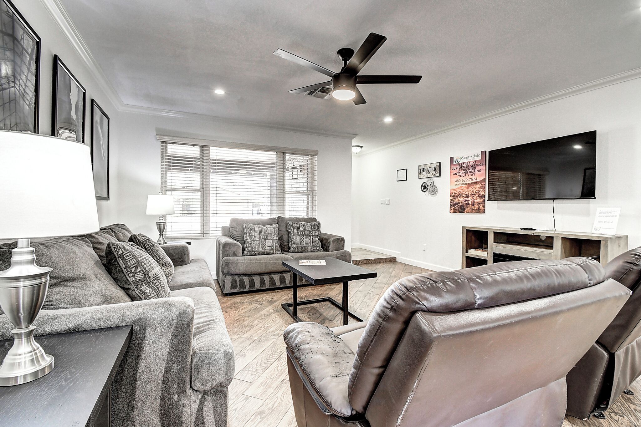 A modern living room with a sofa, armchair, and coffee table, a ceiling fan, and a flat-screen TV mounted on the wall.