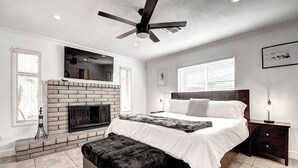 Un dormitorio con chimenea de ladrillo, cama con ropa de cama blanca, una banca, mesita de noche con lámpara y ventilador de techo.