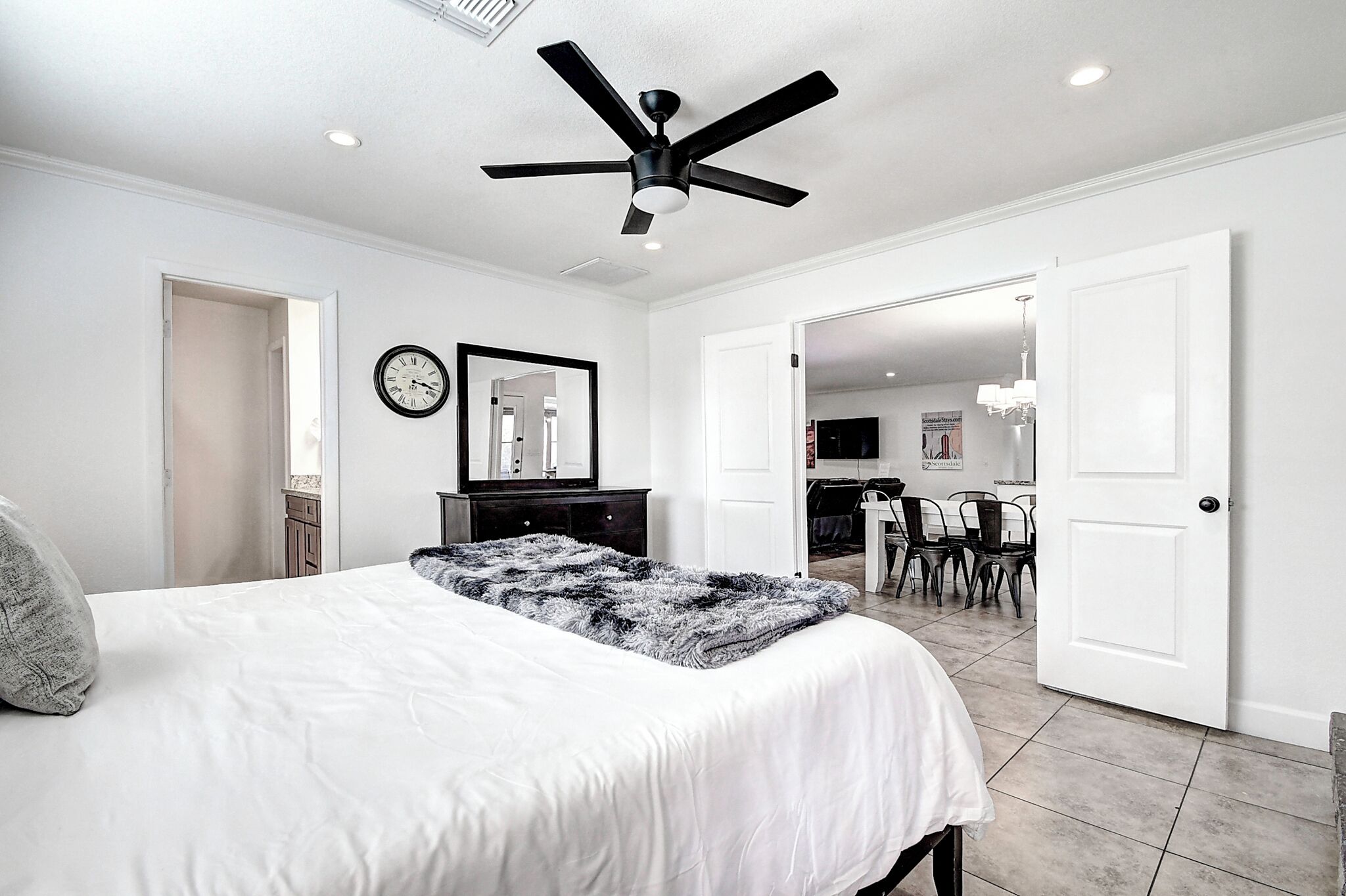 A bedroom with a bed, a ceiling fan, a clock, and a door leading to a dining area.