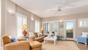 A spacious living room with a beige wall, a ceiling fan, and a large window with white shutters.