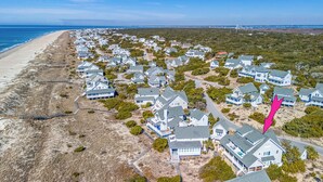 An aerial view of a coastal residential area with houses aligned along the beach, featuring a mix of single and multi-story buildings, surrounded by vegetation and a clear sky.
