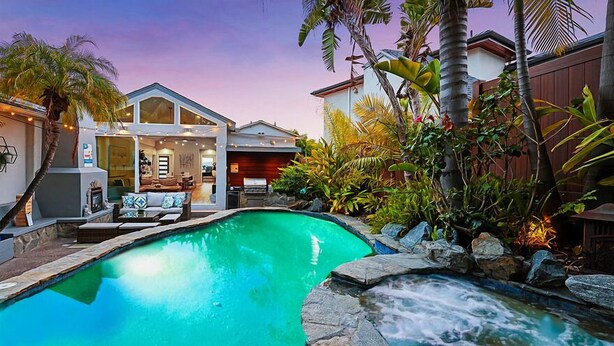 A swimming pool with a waterfall feature, surrounded by tropical plants and a modern house with a visible interior.