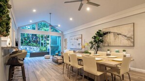 A modern dining area with a large table and chairs, a ceiling fan, and a view of a lush garden through large windows.