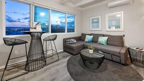 A modern living room with a grey sofa, a round coffee table, and a view of the cityscape through large windows. - Modern Studio - Stunning Views! Centrally Located! (San Diego)
