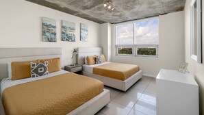 A modern bedroom with two beds, a nightstand, a desk, and a large window with a city view.
