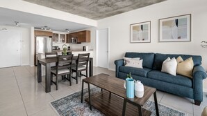 A modern living room with a blue sofa, wooden dining table, and a kitchen in the background.