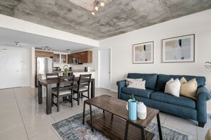 A modern living room with a blue sofa, wooden dining table, and a kitchen in the background.
