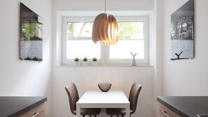 A modern kitchen with a white table, two chairs, a hanging pendant light, and a window with plants.