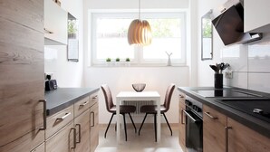 A modern kitchen with wooden cabinets, a dining table with chairs, a pendant light, and a window with blinds.