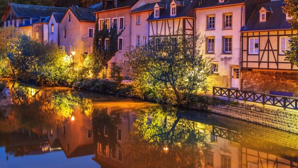 A riverside town with traditional European architecture, including half-timbered houses and a castle in the background.