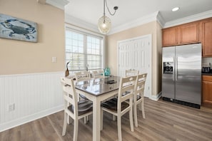 Un área de comedor con mesa y sillas de madera, una lámpara colgante, y una cocina con armarios de madera y un refrigerador de acero inoxidable.