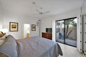 Une chambre avec un lit, une commode, un téléviseur et une porte-fenêtre coulissante donnant sur une terrasse équipée d’une chaise et d’un palmier.