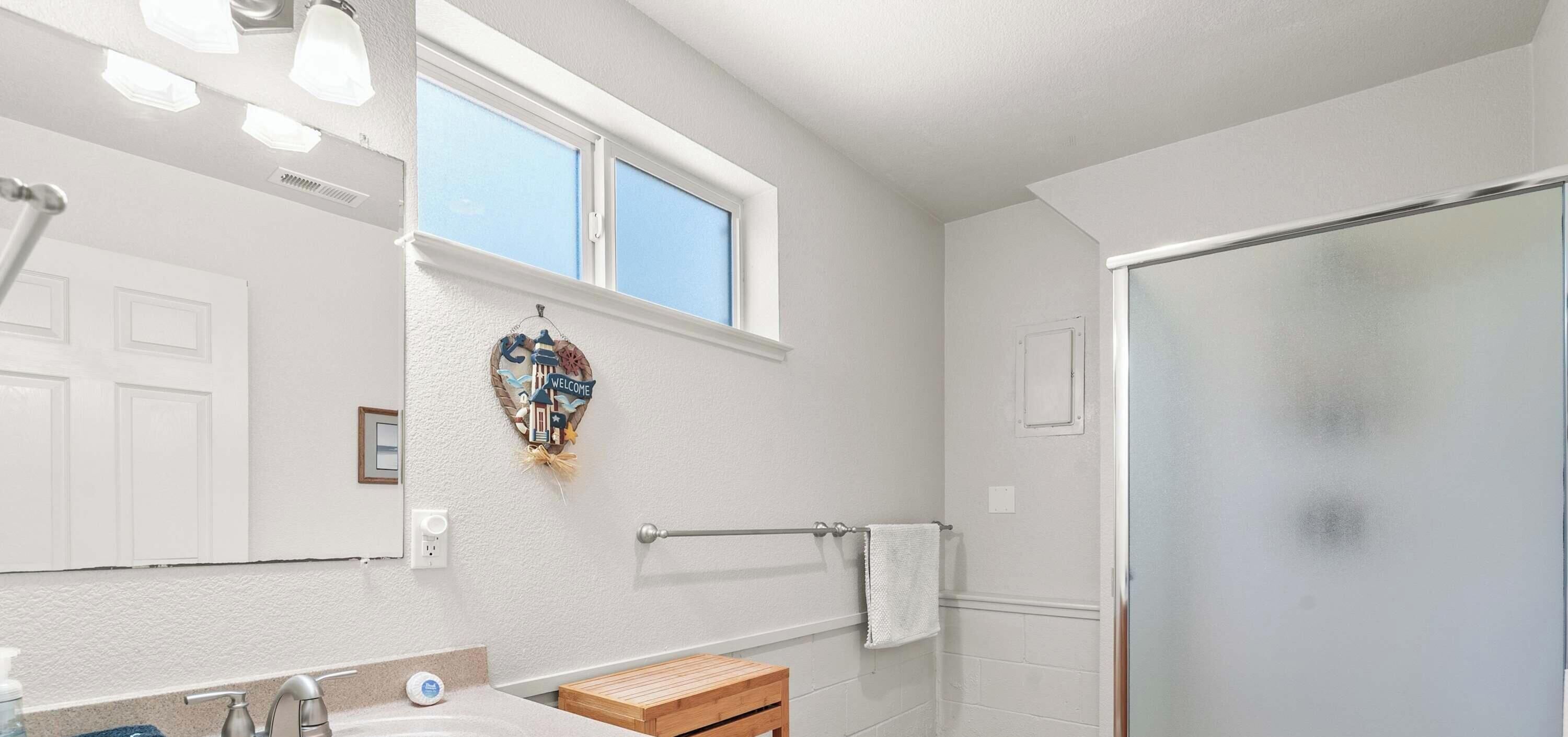 A bathroom with a shower, a towel rack, a window, and a decorative welcome sign.
