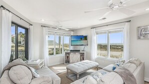 A spacious living room with a large flat-screen TV, comfortable seating, and a view of the beach.