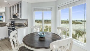 A kitchen with white cabinets, a dark wooden table, and chairs. There is a view of the beach through large windows.