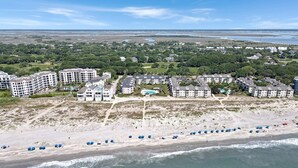 An aerial view of a coastal area with beachfront properties, including multi-story buildings and a sandy beach with umbrellas.