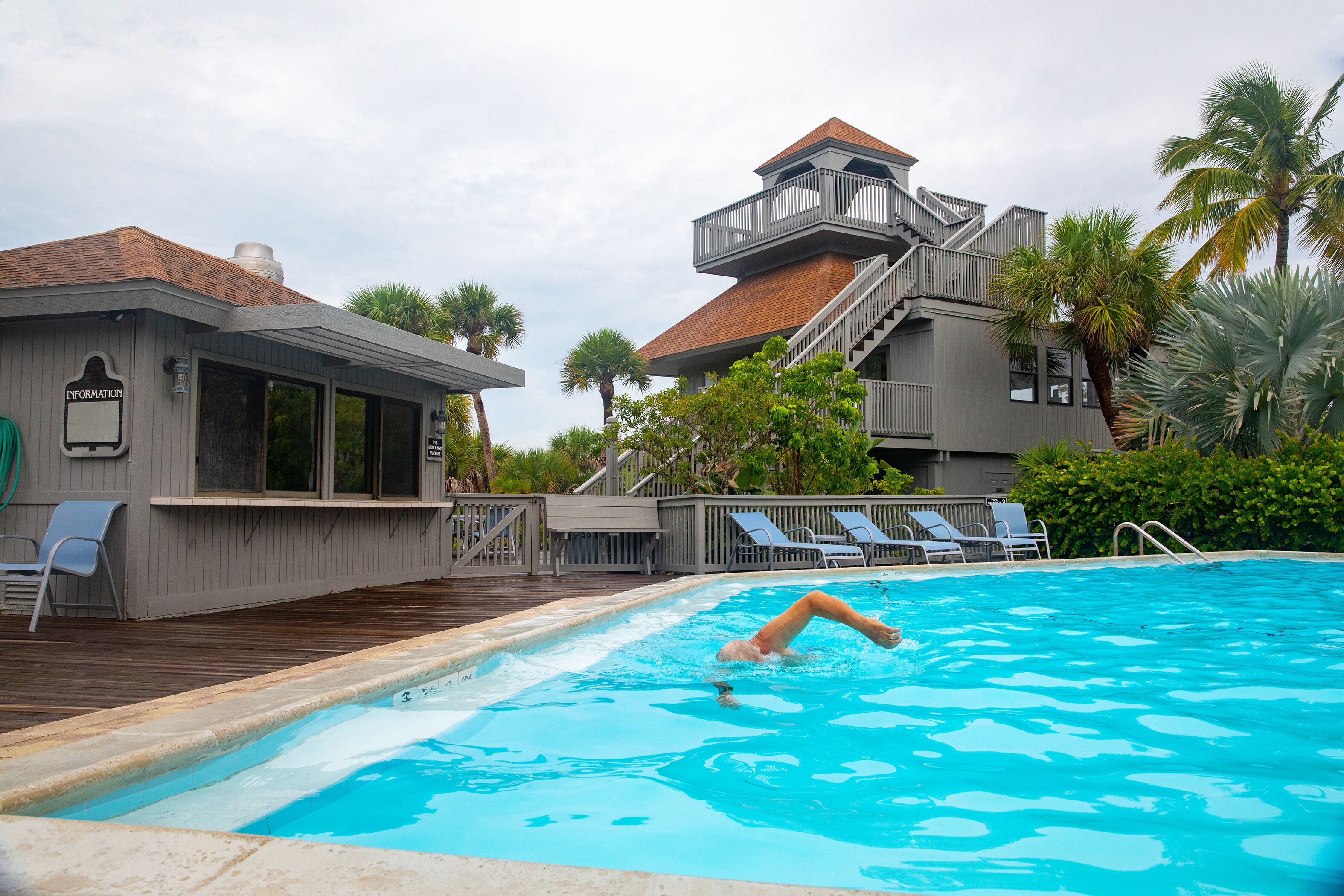 En swimmingpool med en person som simmar, omgiven av ett poolområde med solstolar och en byggnad med en balkong.