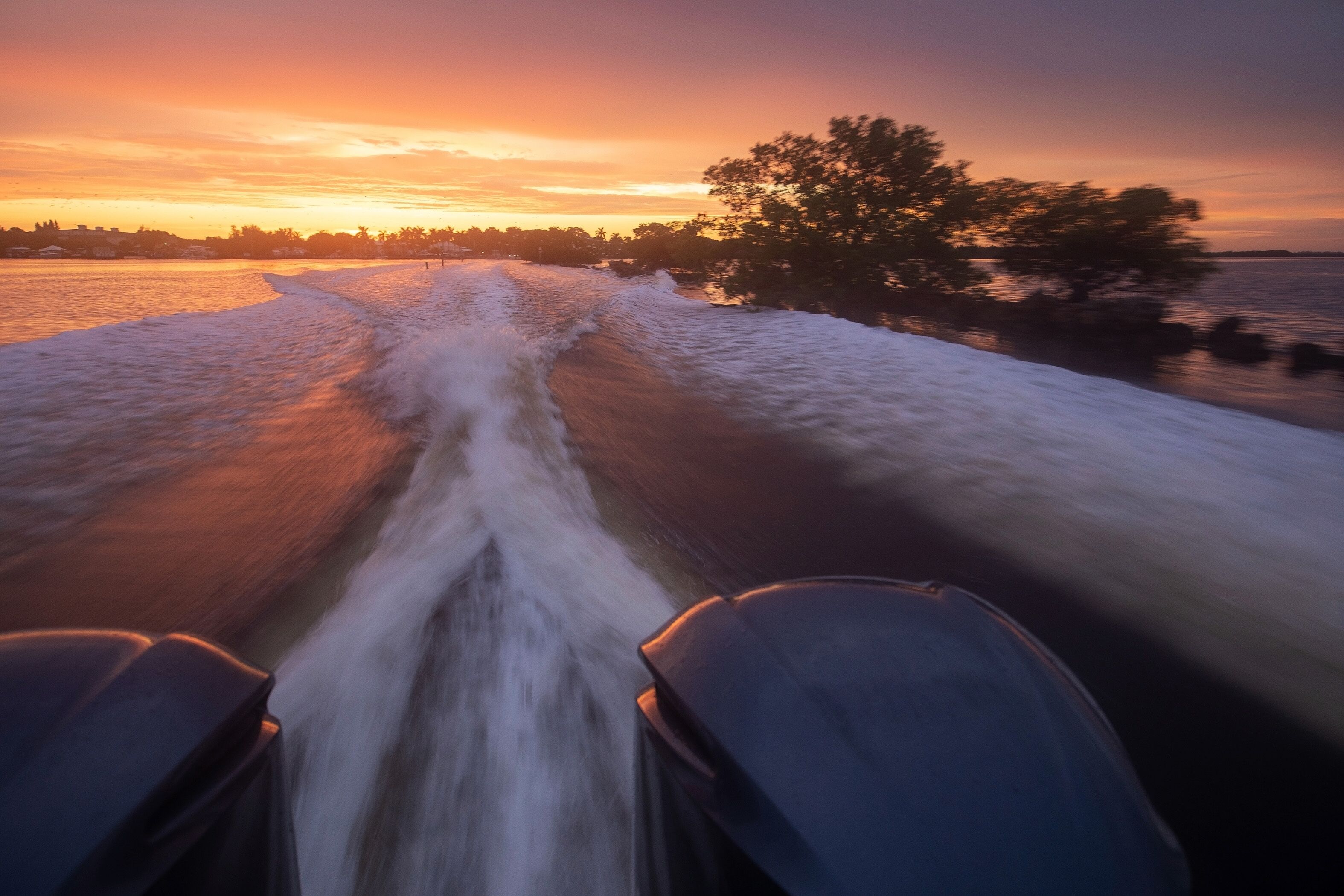 Une rivière où un bateau navigue, laissant une vague derrière lui, le tout entouré d’arbres et illuminé par un coucher de soleil.