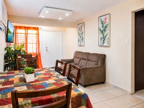 A living room with a sofa, a dining table with chairs, a TV, and a door with curtains. - Jobos Beach Apartment A-3 (Isabela)