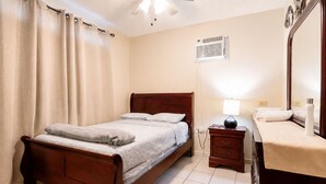 A bedroom with a wooden bed, a bedside table with a lamp, a mirror, and a window with blinds. - Jobos Beach Apartment A-3 (Isabela)