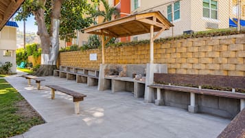 Un área de estar al aire libre con bancos de hormigón y una pérgola de madera, rodeada por un muro de ladrillo y un árbol.