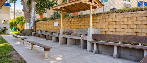 Un área de estar al aire libre con bancos de hormigón y una pérgola de madera, rodeada por un muro de ladrillo y un árbol.