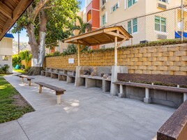 Un área de estar al aire libre con bancos de hormigón y una pérgola de madera, rodeada por un muro de ladrillo y un árbol.
