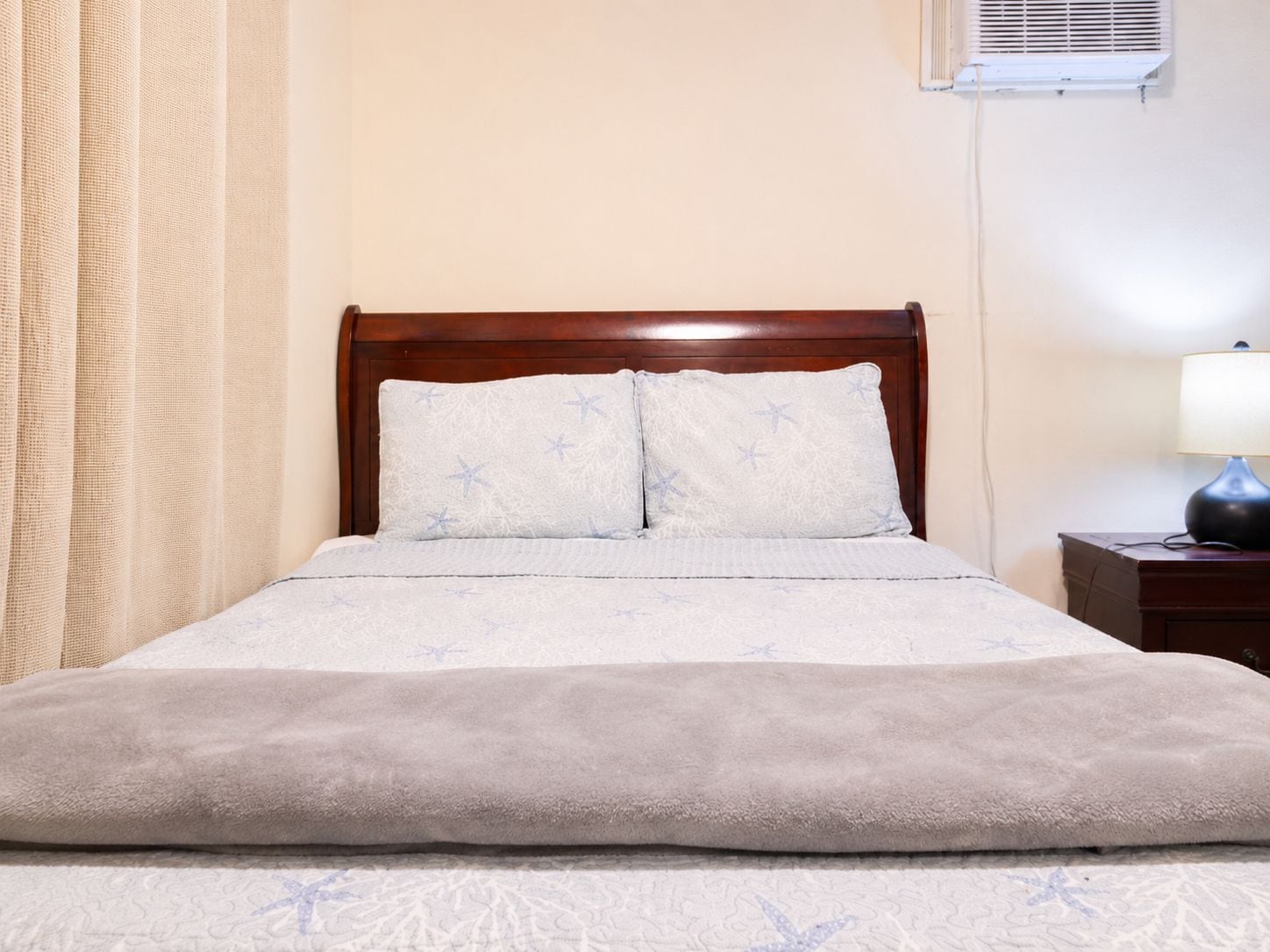 Una cama con cabecera, dos almohadas, una manta y una mesita de noche con lámpara.
