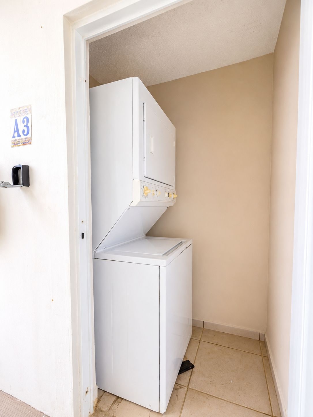 Uma lavanderia compacta e branca, equipada com uma máquina de lavar e uma secadora, uma porta com a placa 'A3' e chão revestido de azulejos.