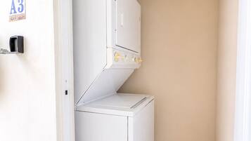 Uma lavanderia compacta e branca, equipada com uma máquina de lavar e uma secadora, uma porta com a placa 'A3' e chão revestido de azulejos.